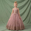 A young girl with blonde hair in a bun stands wearing a sleeveless dusty pink ball gown with gold floral embroidery and a tulle skirt, posed against a draped sage green backdrop.