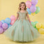 A young girl stands in a studio wearing an off-the-shoulder sage green tulle ball gown with gold embroidered details. The background is a soft yellow with pastel balloons in pink, blue, purple, green, and yellow.