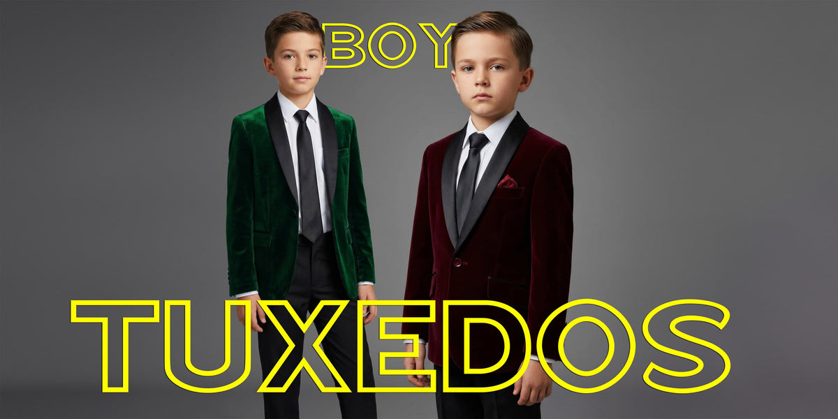 Two young boys modeling formal tuxedos against a dark gray studio background. The boy on the left wears a emerald green velvet tuxedo jacket with a black tie and white shirt.