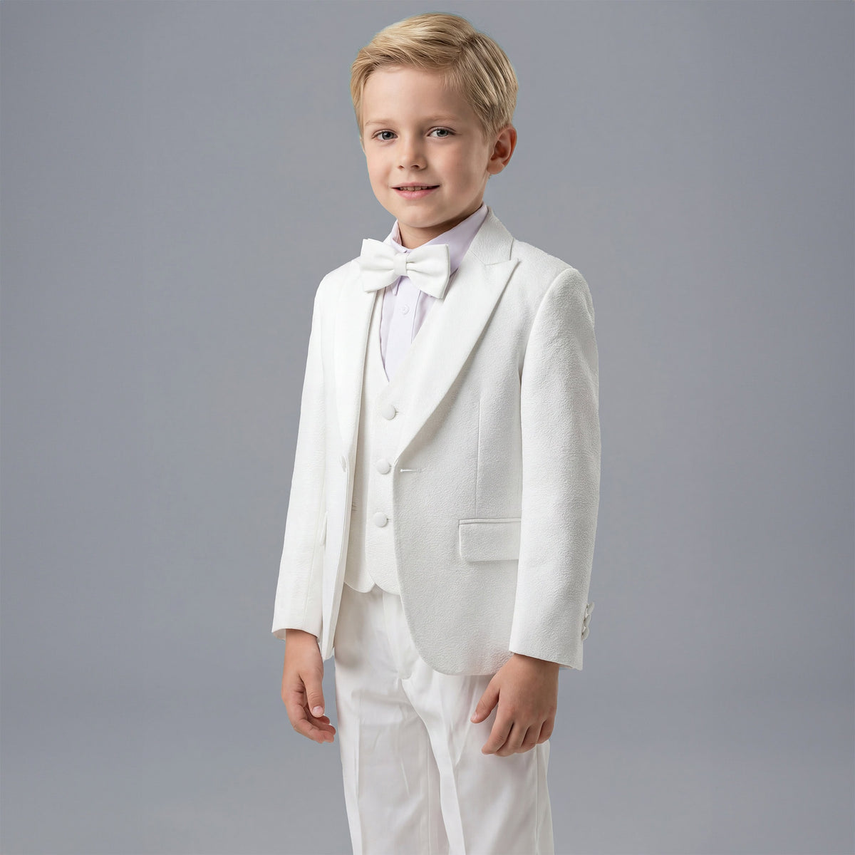 A young boy modeling a white formal kids' suit consisting of a jacket, vest, trousers, white shirt, and a matching bow tie, set against a solid gray studio background. Main image
