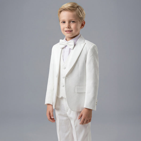 A young boy modeling a white formal kids' suit consisting of a jacket, vest, trousers, white shirt, and a matching bow tie, set against a solid gray studio background.