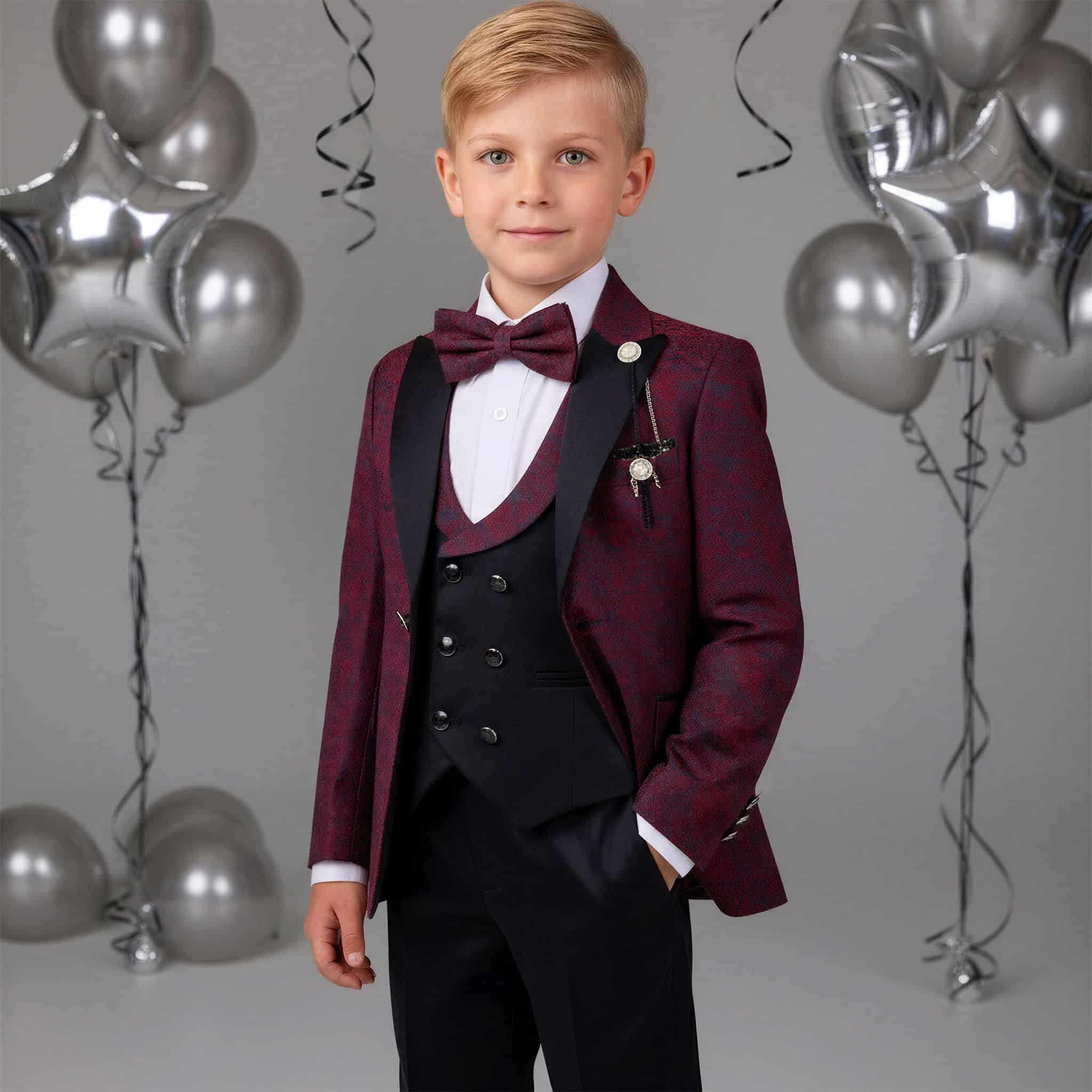 A young boy modeling a formal kids' tuxedo with a burgundy patterned jacket, black vest and trousers, a matching burgundy bow tie, and a white dress shirt. The background is a gray studio setting decorated with silver balloons. Main image