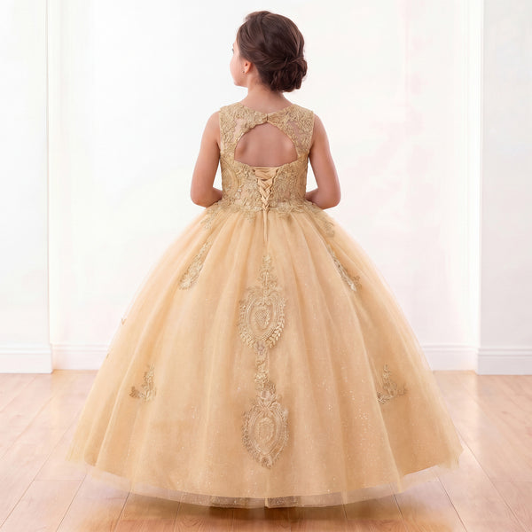 A young girl viewed from behind wearing a voluminous champagne-gold ball gown with intricate lace and tulle embroidery, standing on a light wood floor against a white wall.