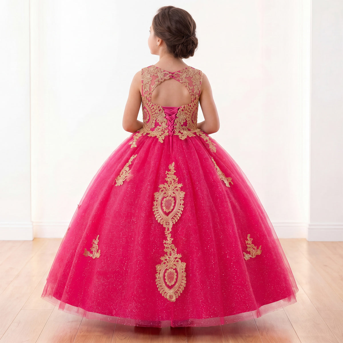 A young girl viewed from behind wearing a bright pink ball gown with gold lace embroidery and a glittery tulle skirt, standing in a bright indoor room with light wood floors.
