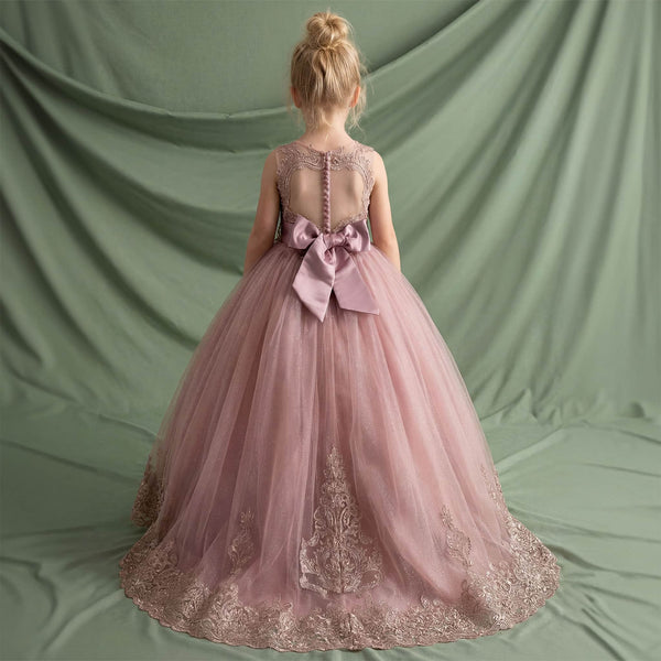 A young girl viewed from behind wearing a dusty pink ball gown with a large satin bow at the back, lace detailing, and a full tulle skirt, standing against a draped sage green fabric backdrop.