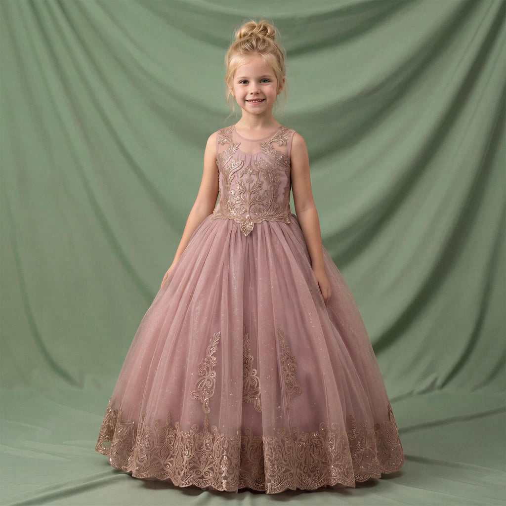A young girl with blonde hair in a bun stands wearing a sleeveless dusty pink ball gown with gold floral embroidery and a tulle skirt, posed against a draped sage green backdrop.