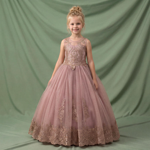 A young girl with blonde hair in a bun stands wearing a sleeveless dusty pink ball gown with gold floral embroidery and a tulle skirt, posed against a draped sage green backdrop.