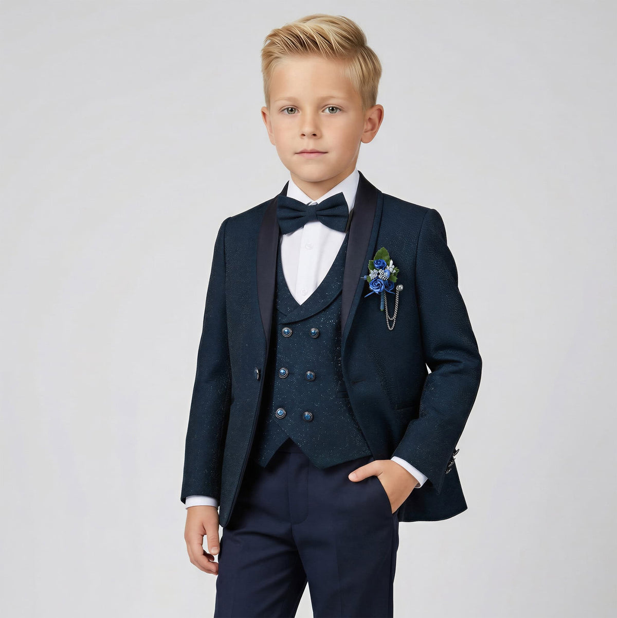 A young boy modeling a dark navy blue three-piece formal suit with a matching bow tie and a blue floral boutonniere, set against a plain light gray studio background. Main image