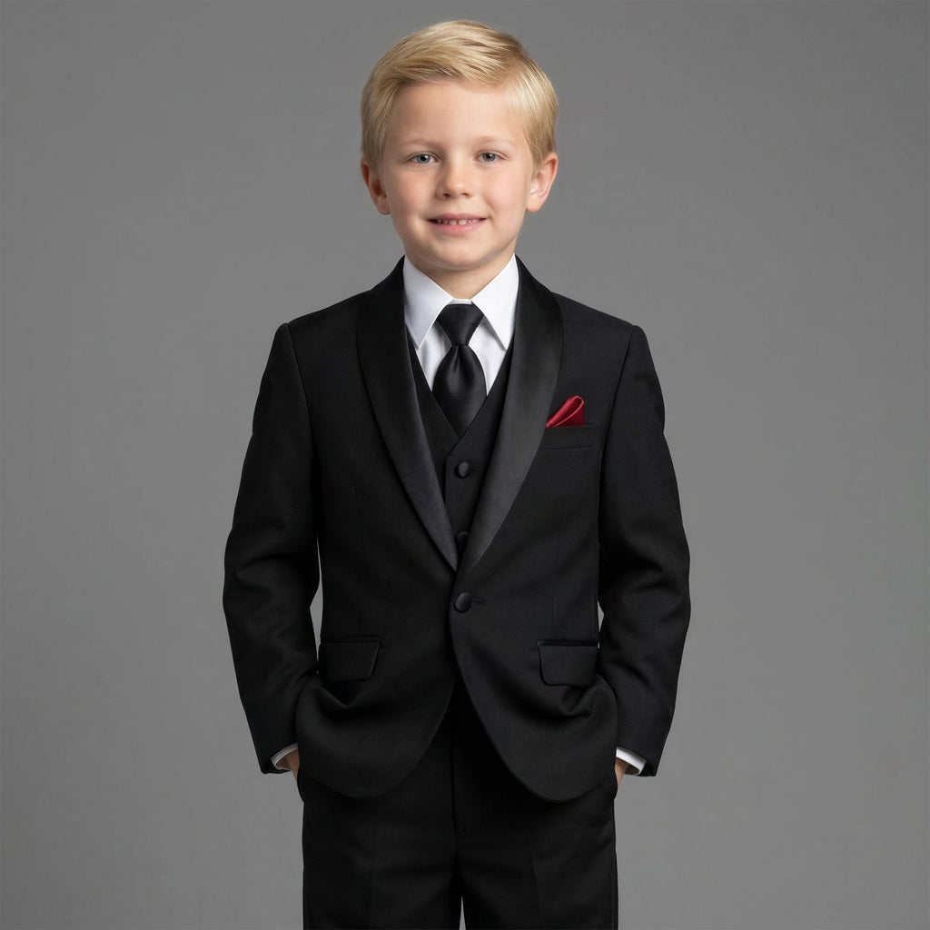 A young blond boy wearing a black formal suit with black satin lapels, a white dress shirt, a black tie, and a red pocket square, standing against a solid gray studio background.