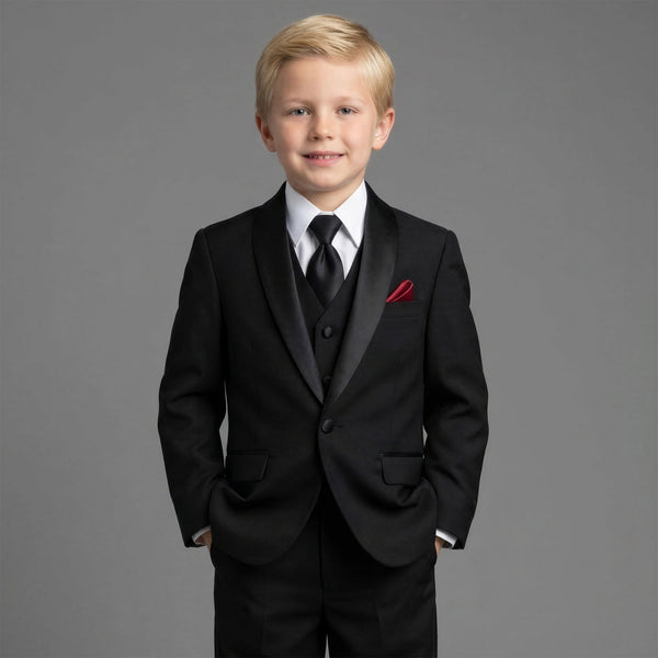 A young blond boy wearing a black formal suit with black satin lapels, a white dress shirt, a black tie, and a red pocket square, standing against a solid gray studio background.