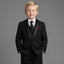A young blond boy wearing a black formal suit with black satin lapels, a white dress shirt, a black tie, and a red pocket square, standing against a solid gray studio background.