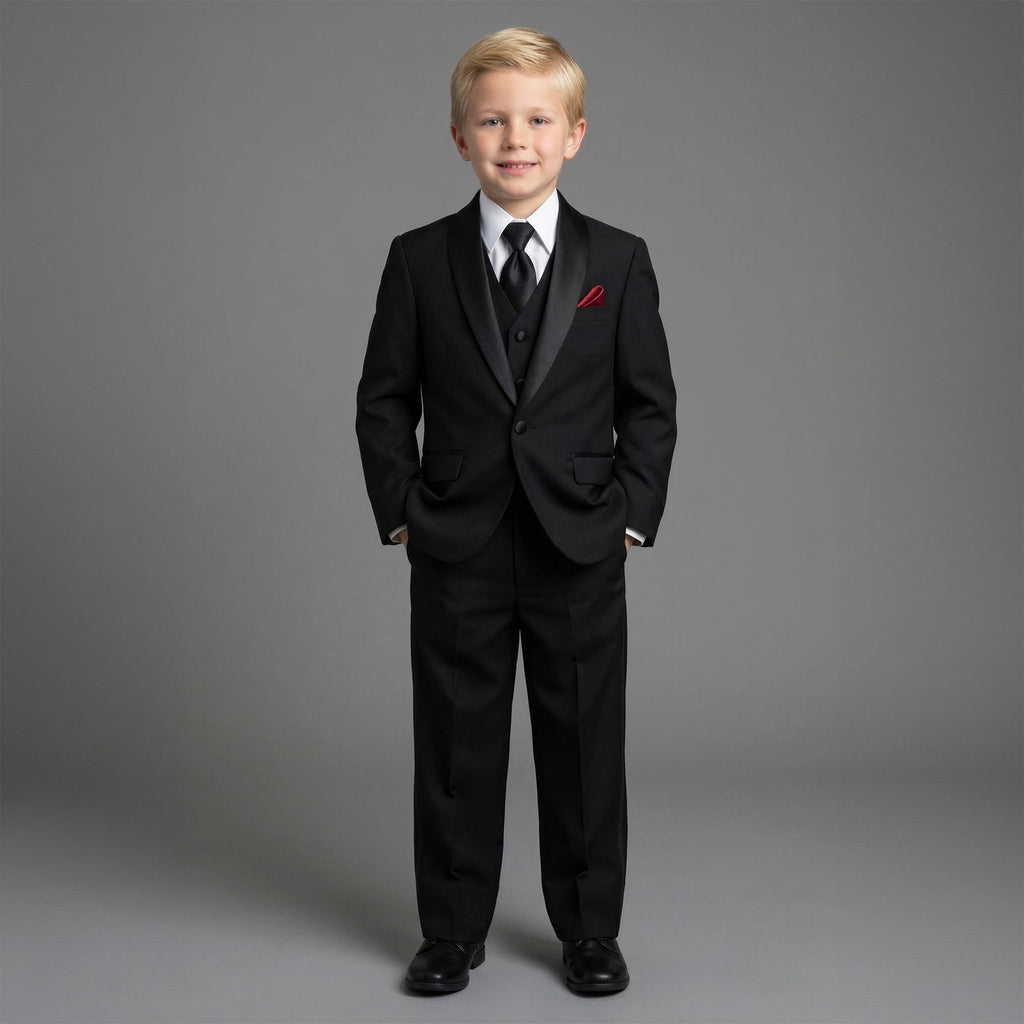 A young boy modeling a black three-piece formal suit with satin lapels, a white dress shirt, a black tie, and a red pocket square, standing against a solid gray studio background.