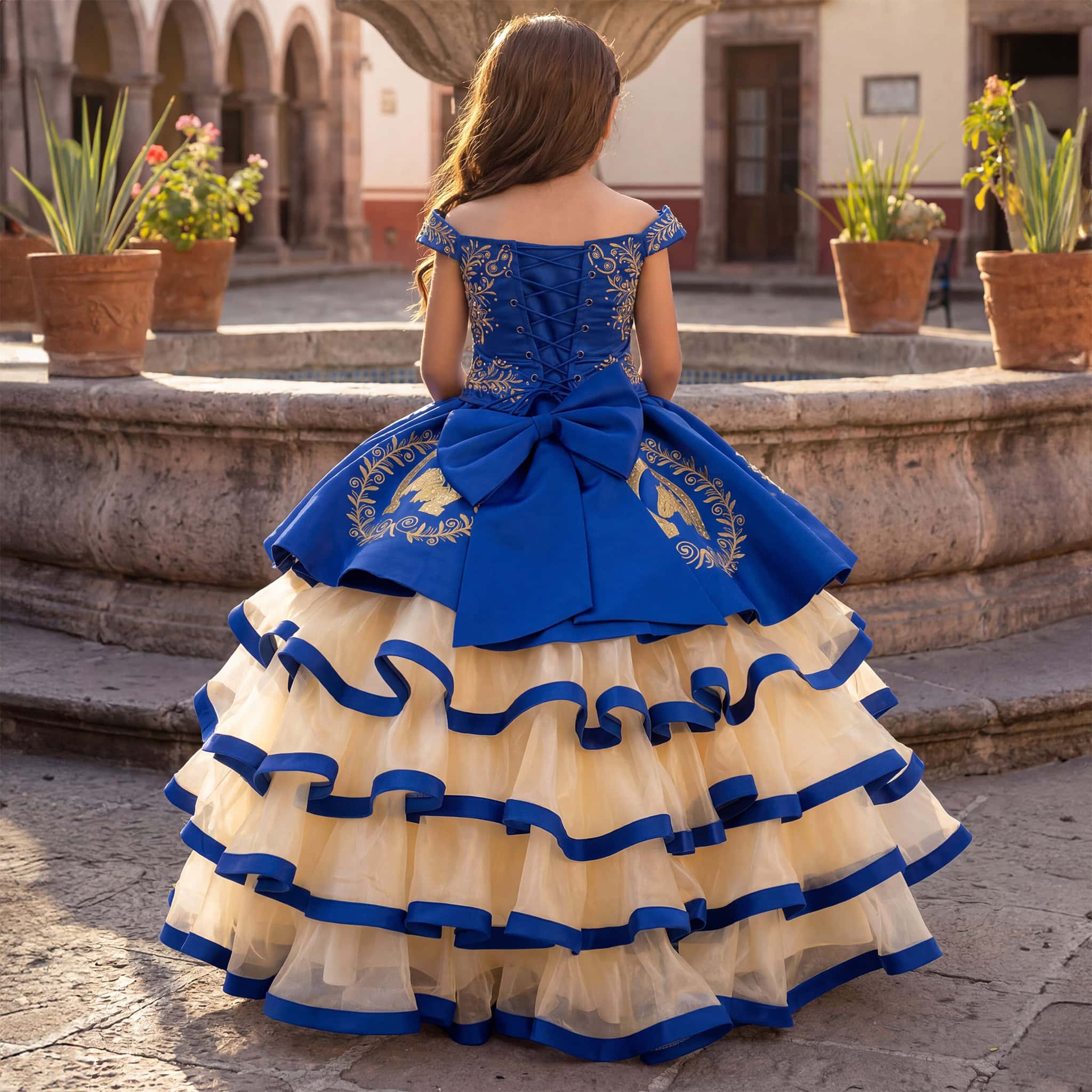 A young girl viewed from behind wearing a royal blue and cream tulle ball gown quinceañera dress, standing in front of a stone courtyard fountain.