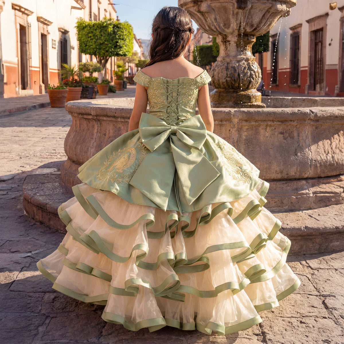 A young girl viewed from behind wearing an elaborate off-the-shoulder ball gown in a cobblestone plaza with a stone fountain in the background. The dress features a sage green satin bodice with a large bow, gold embroidery, and a layered tulle skirt with ruffled trim.