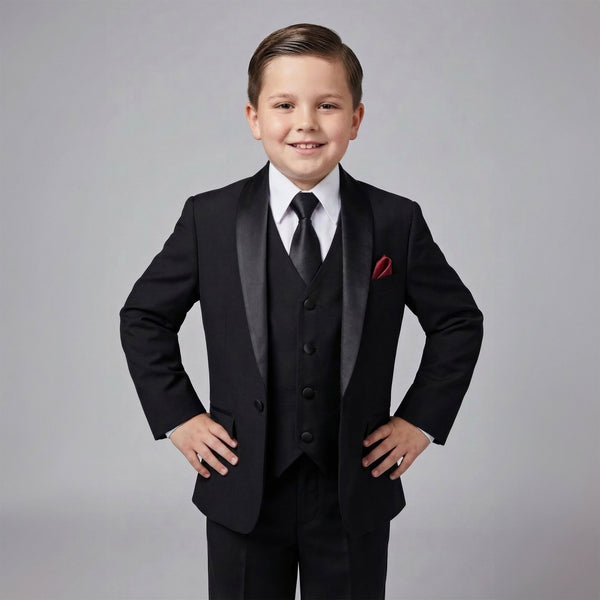 A young boy modeling a black formal three-piece suit with satin lapels, a white dress shirt, a black tie, and a red pocket square, posed against a solid gray studio background.