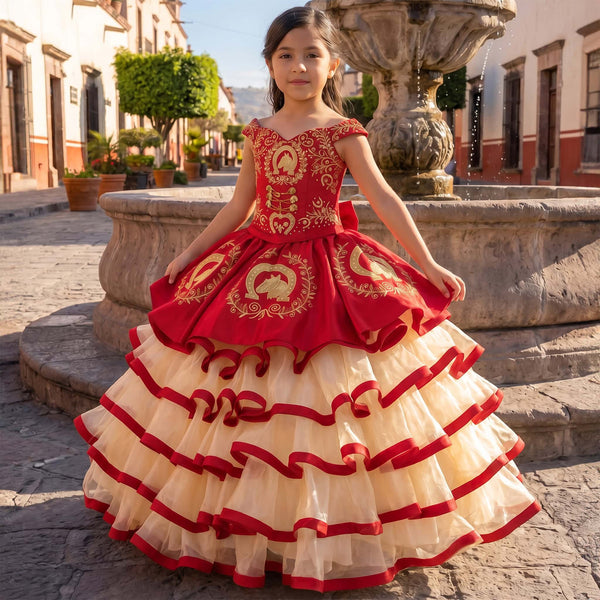 A young girl wearing a traditional Mexican red and cream tulle ball gown stands in front of a stone fountain in a historic town square.