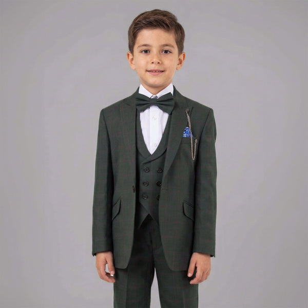 A young boy modeling a dark green three-piece suit with a matching bow tie, white dress shirt, and a blue pocket square against a solid gray studio background.