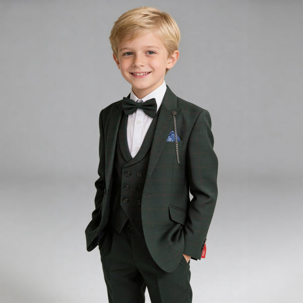 A young blond boy modeling a dark green three-piece suit with a subtle plaid pattern, featuring a matching vest, trousers, black bow tie, and a blue patterned pocket square, set against a solid gray studio background.