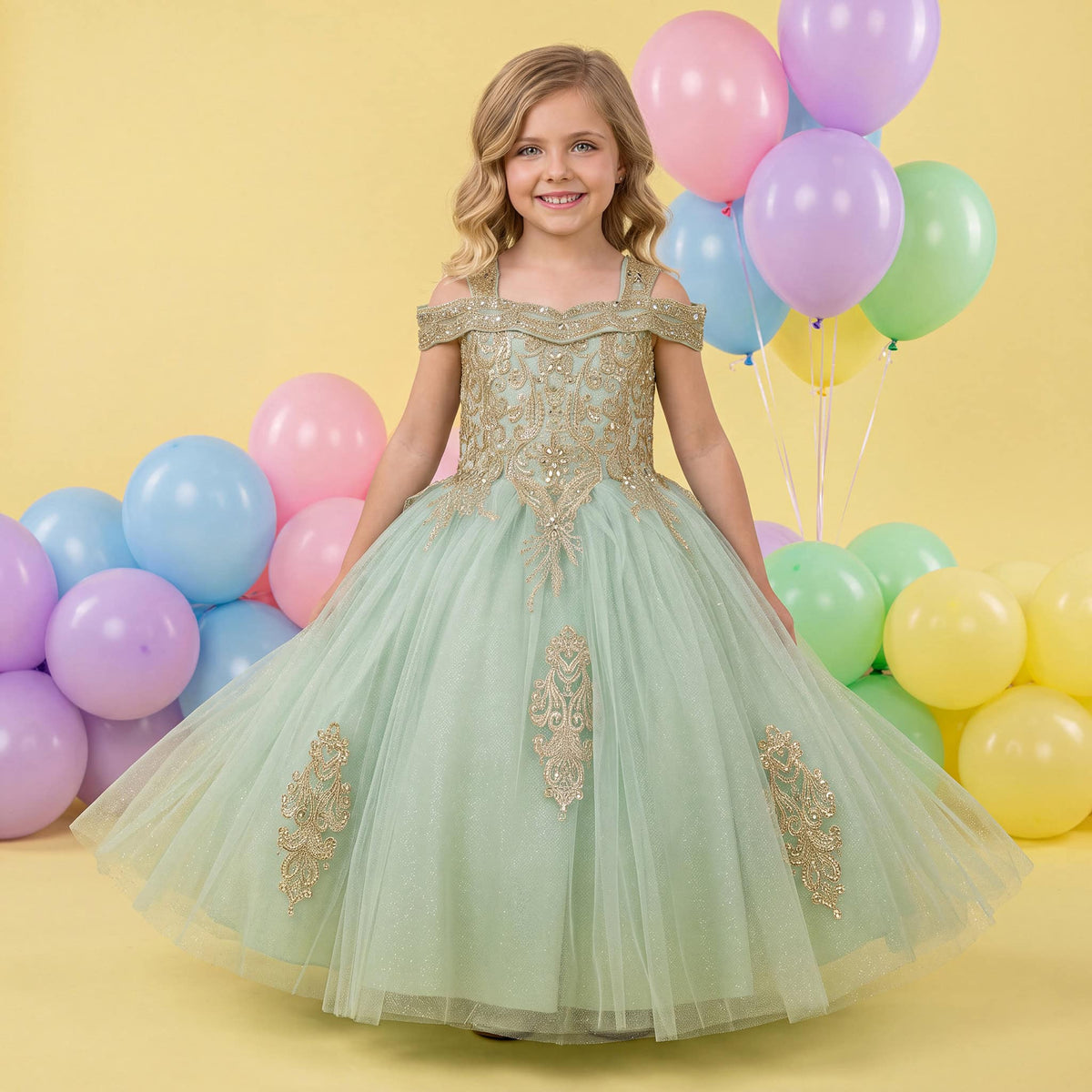 A young girl stands in a studio wearing an off-the-shoulder sage green tulle ball gown with gold embroidered details. The background is a soft yellow with pastel balloons in pink, blue, purple, green, and yellow. Main image