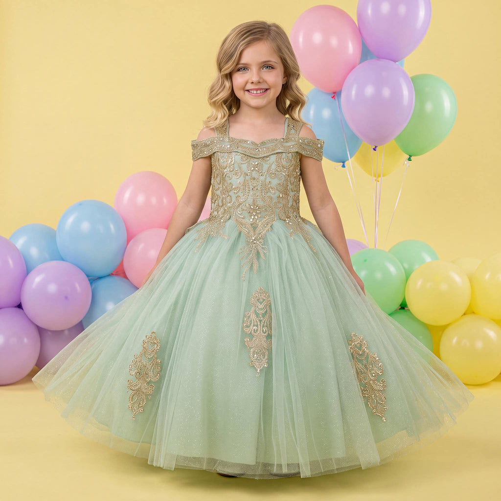 A young girl stands in a studio wearing an off-the-shoulder sage green tulle ball gown with gold embroidered details. The background is a soft yellow with pastel balloons in pink, blue, purple, green, and yellow.