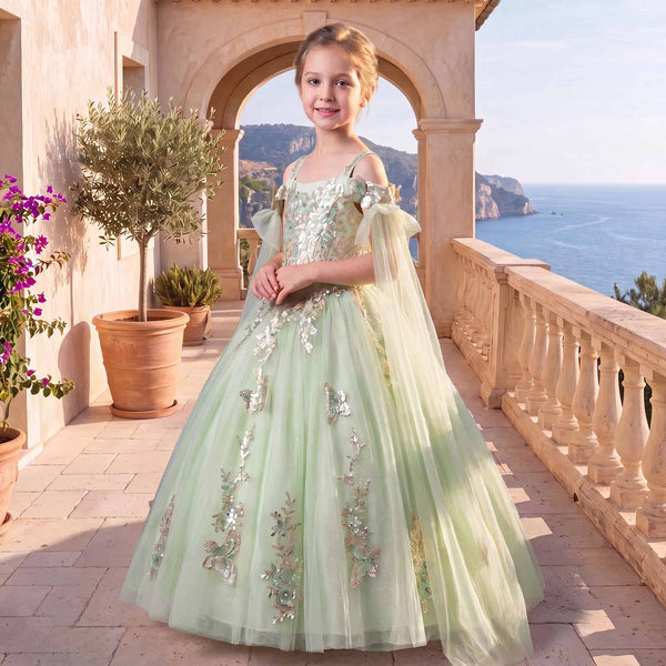 A young girl stands on a sunlit stone terrace wearing a pale mint green ball gown with silver floral embroidery and sheer bow details. The background features an archway, potted plants, and a scenic view of the sea.